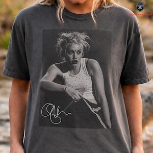 Peut inclure: T-shirt gris fonc&eacute; avec une image en noir et blanc d'une femme portant un d&eacute;bardeur blanc &agrave; pois. L'image comprend une signature. Le t-shirt a des manches courtes.