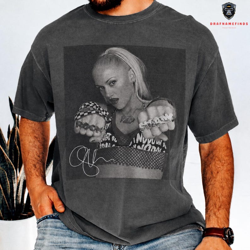 Peut inclure: T-shirt gris fonc&eacute; avec une image en noir et blanc d'une femme blonde attach&eacute;e en queue de cheval. L'image comprend le texte "ROCK" et "STARS" sur les bagues, et une signature. Le t-shirt a des manches courtes.