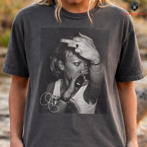 Peut inclure: T-shirt gris fonc&eacute; avec une image en noir et blanc d'une personne tenant un microphone et faisant un doigt d'honneur. L'image comprend une signature blanche. Le t-shirt est de style vintage.