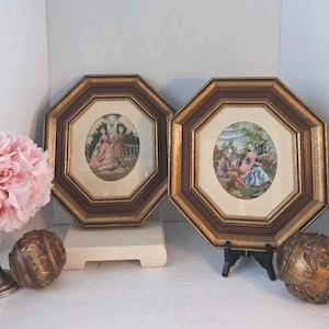 May include: Two octagonal framed prints featuring oval-shaped illustrations of figures in period clothing. The frames are dark brown and gold with cream-coloured matting. Each print depicts a scene with people in a garden setting.