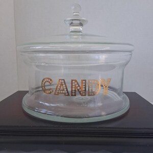 Könnte beinhalten: Ein durchsichtiges Glasgefäß mit passendem Deckel und einem knopfförmigen Griff. Das Wort CANDY ist in metallisch goldenen Buchstaben auf der Vorderseite des Glases aufgedruckt, wobei jeder Buchstabe ein dekoratives Punktmuster aufweist.