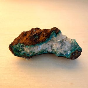 May include: A natural mineral specimen featuring a rough, brown exterior with vibrant turquoise and teal accents. The center reveals a cluster of clear, crystalline formations, creating a striking contrast in color and texture. The specimen is displayed on a light-colored surface.