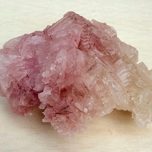 Peut inclure: Spécimen minéral cristallin avec une structure en couches. La formation rocheuse présente un dégradé de couleurs, passant d'un rose profond à une teinte pâle, presque translucide. La surface est rugueuse et texturée.