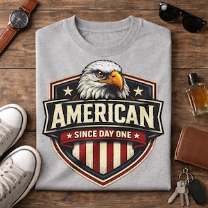 May include: A gray t-shirt featuring a graphic of an American bald eagle with the text "AMERICAN SINCE DAY ONE" in a shield design. The eagle has white feathers, an orange beak, and a stern expression.