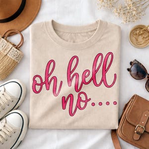 Könnte beinhalten: Beiges Sweatshirt mit Rundhalsausschnitt und dem Schriftzug "oh hell no..." in rosa. Das Sweatshirt ist sauber auf einer weißen Oberfläche gefaltet, mit Accessoires wie Sonnenbrille und Tasche im Hintergrund.