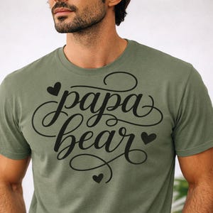 May include: Olive green t-shirt with the words "papa bear" in a black, cursive font. The text is adorned with three small black hearts. The shirt is made of a soft material.