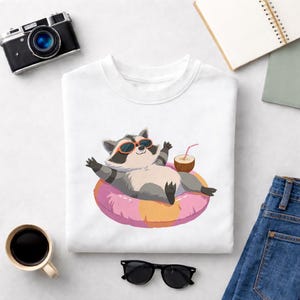 Op de afbeelding: Wit sweatshirt met een cartoon wasbeer met een zonnebril, ontspannen op een roze en oranje opblaasbare ring. De wasbeer houdt een kokosdrankje vast. Een zwarte camera, zonnebril en een kopje koffie zijn ook zichtbaar.