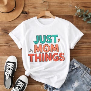 Może przedstawiać: Biała koszulka z napisem "JUST MOM THINGS" w kolorze turkusowym, koralowym i czerwonym. Projekt zawiera filiżankę kawy i symbol serca. Koszulka jest prezentowana z czarno-białymi trampkami i szortami dżinsowymi na drewnianej powierzchni.