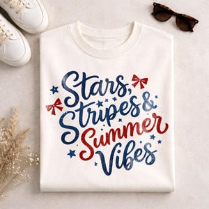 以下が含まれることがあります： 白地のTシャツに「Stars, Stripes & Summer Vibes」の文字が青と赤で書かれています。デザインには星とリボンのアクセントが含まれています。Tシャツは折りたたまれ、白いスニーカーとサングラスと一緒に表示されています。