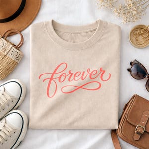 May include: A beige crew neck t-shirt with the word "Forever" in coral script. The shirt is folded and surrounded by accessories, including a brown hat, purse, sunglasses, and trainers.