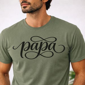 May include: Olive green t-shirt with the word "papa" in black script. The word is written in a decorative font with flourishes. The shirt has short sleeves and a crew neck.