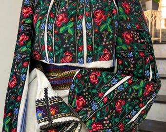 May include: A traditional embroidered blouse with a black background, featuring red roses and blue floral accents. The sleeves and bodice are intricately detailed with colorful stitching. The blouse is paired with a white skirt and a colorful belt.