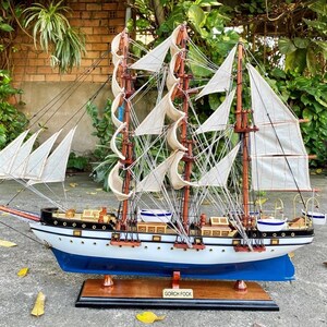 Puede incluir: Modelo detallado de un velero con velas blancas, mástiles marrones y casco azul. El barco está montado sobre una base de madera, y el nombre "GORCH FOCK" es visible en una pequeña placa. El modelo se encuentra sobre un fondo de follaje verde.