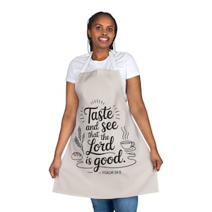 May include: Beige apron with the text "Taste and see that the Lord is good. Psalm 34:8" in black script. The apron features illustrations of wheat, bread, and a cup of coffee. The apron has a white neck strap.