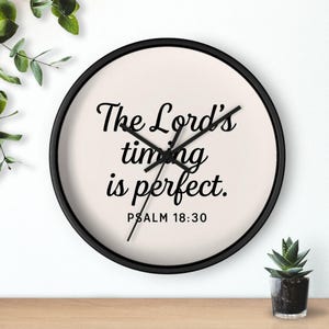 Op de afbeelding: Een ronde wandklok met een zwart frame en een crèmekleurige wijzerplaat. De klok toont de tekst "The Lord's timing is perfect. Psalm 18:30" in zwart schrift. De wijzers van de klok zijn zwart.