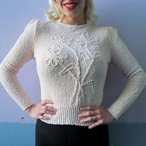 Puede incluir: Un suéter de punto color crema con un diseño floral. El suéter tiene mangas largas y un cuello redondo.