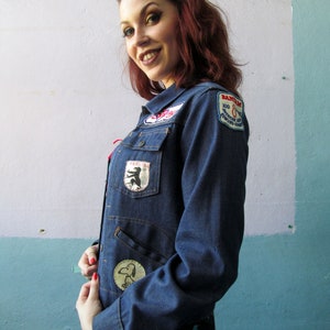 May include: A denim jacket with various patches, including a Snoopy patch and a patch that says "Bantam 100". The jacket has a classic design with a button-up front and pockets. The jacket is worn over a floral print garment.