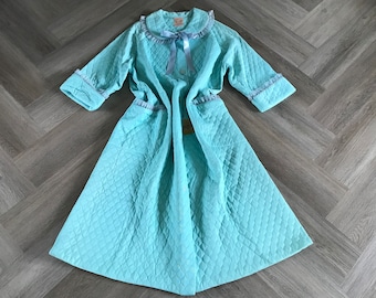 May include: A light blue quilted robe with a diamond pattern. The robe has a ruffled collar and cuffs, with a light blue ribbon and button closure. The robe is laid out on a light gray wooden surface.