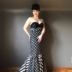 Puede incluir: Un top negro sin tirantes combinado con una falda larga de cintura alta. La falda presenta lunares en la parte superior y rayas blancas y negras en la sección inferior acampanada. El conjunto es de diseño de inspiración vintage.