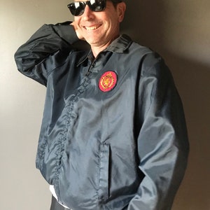 May include: A dark gray nylon jacket with a red and gold emblem on the chest. The jacket has a zipper closure, a collar, and a pocket on the front. The person in the image is wearing black sunglasses.