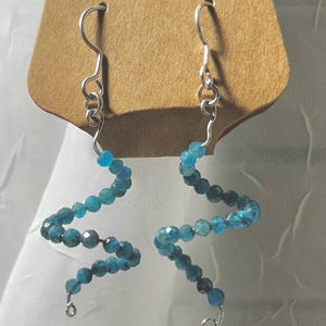 Puede incluir: Pendientes hechos a mano con un diseño ondulado único. Cada pendiente presenta una serie de cuentas facetadas de color verde azulado ensartadas en alambre plateado. Los pendientes se exhiben en una etiqueta marrón con el texto "Handmade with love" y un corazón rojo.
