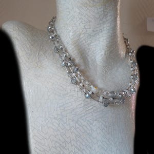 May include: A multi-strand necklace with clear and silver beads and accents. The necklace is displayed on a white mannequin. The necklace has a delicate, elegant design, with a mix of bead sizes and textures.