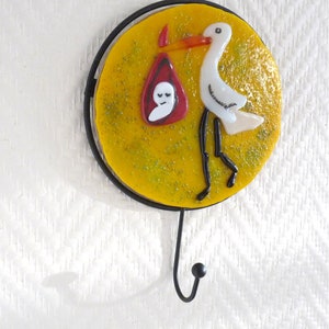 May include: A yellow and white glass stork carrying a baby in a red bag, mounted on a black metal hook.