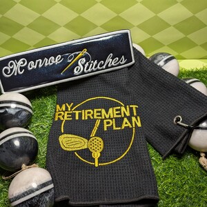 May include: Black golf towel with the text "MY RETIREMENT PLAN" in yellow, along with a golf club and ball graphic. A black patch with "Monroe Stitches" embroidered in white and gold is also present. Decorative golf balls are in the background.