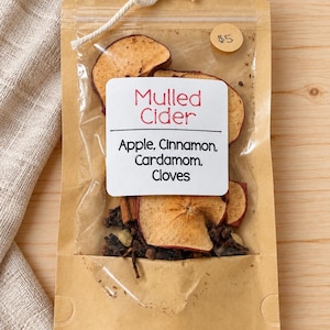 May include: A brown paper pouch with a clear window containing dried apple slices, cinnamon sticks, cardamom pods, and cloves. A white label reads Mulled Cider, Apple, Cinnamon, Cardamom, Cloves. The pouch is tied with a white string at the top.