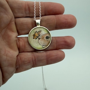May include: A silver-toned pendant necklace featuring a circular charm with an illustration of a small brown animal with a butterfly resting on its nose. The pendant is attached to a delicate silver-colored chain with a standard metal clasp.