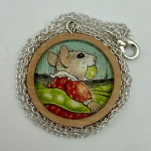 May include: A round pendant necklace featuring a circular wooden frame containing a printed illustration of a mouse wearing a red and yellow patterned garment, holding a green object, attached to a silver-toned metal chain with a lobster clasp.