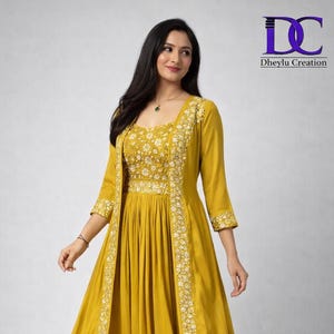 Puede incluir: Vestido largo amarillo mostaza con chaqueta a juego. El vestido y la chaqueta presentan bordados florales blancos en el corpiño y los bordes. El vestido tiene un escote cuadrado y una falda fluida. El logo de la marca "Dheylu Creation" está en la esquina superior derecha.