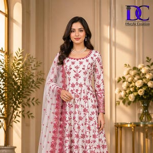 May include: A white, long-sleeved dress with detailed red floral embroidery. The dress has a flowing shape and is worn with a matching sheer scarf. The image also includes the text "DC Dheylu Creation".