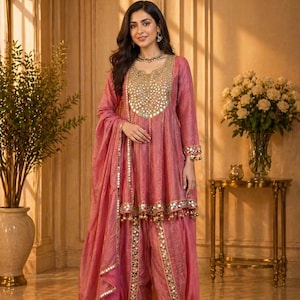 May include: A pink salwar kameez outfit with gold mirror detailing. The set comprises a long-sleeved tunic, matching trousers, and a dupatta. The tunic has an ornate gold neckline. The outfit is accessorized with gold jewellery and heels.