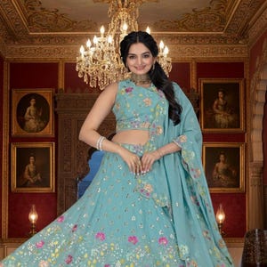Peut inclure: Une robe de cérémonie bleu clair avec broderies florales et ornements de sequins. La robe comprend un haut sans manches et une jupe fluide, assortie d'un châle. La tenue est portée par une personne dans une pièce avec des portraits et un lustre.
