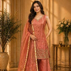 Peut inclure: Ensemble salwar kameez pêche sans manches. Le haut présente des broderies dorées et un col en V. Le pantalon palazzo assorti et le dupatta transparent sont bordés d'or. La tenue est portée avec des chaussures dorées.
