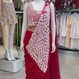 May include: A deep red saree featuring a sleeveless blouse with intricate white geometric embroidery and a matching white peacock feather pattern cascading down the front. The skirt portion has soft, vertical pleats in a solid red fabric.