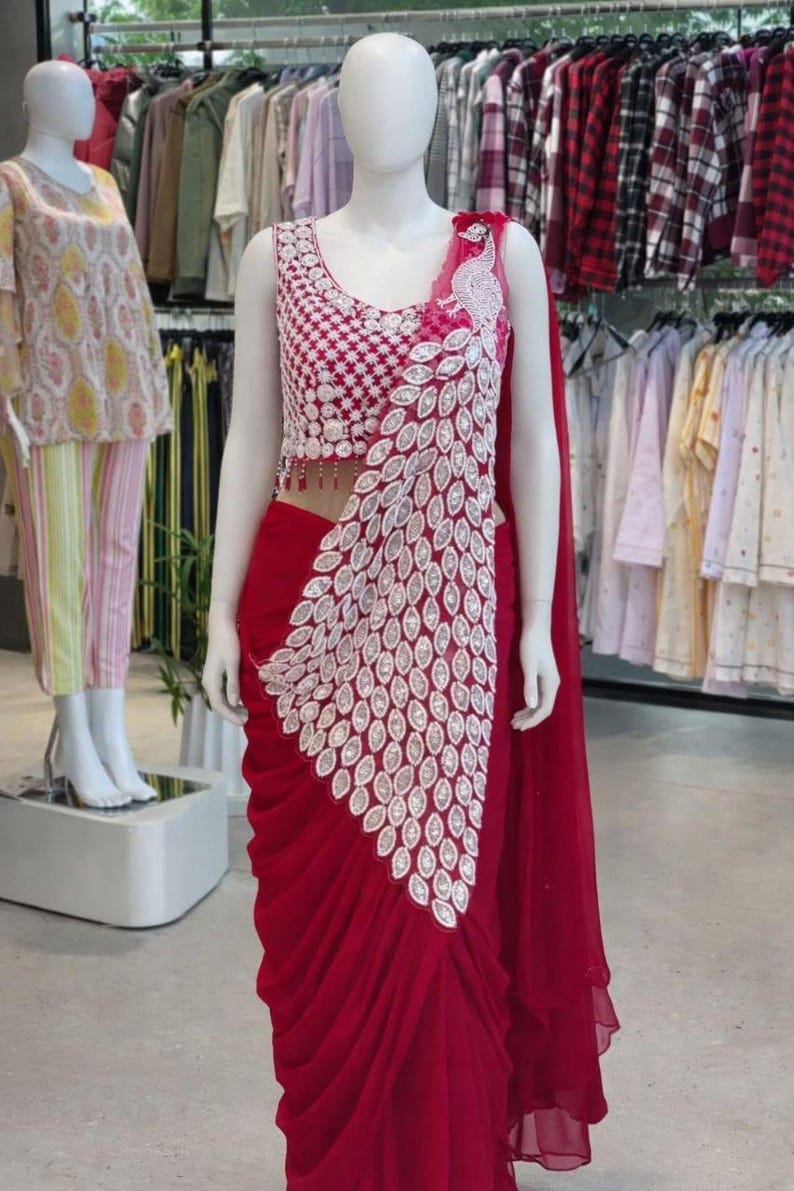 May include: A deep red saree featuring a sleeveless blouse with intricate white geometric embroidery and a matching white peacock feather pattern cascading down the front. The skirt portion has soft, vertical pleats in a solid red fabric.
