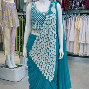 May include: A teal saree featuring a sleeveless blouse with intricate silver embroidery and a matching teal skirt with a prominent silver peacock feather pattern draped across the front. The ensemble includes a long, flowing teal fabric panel.
