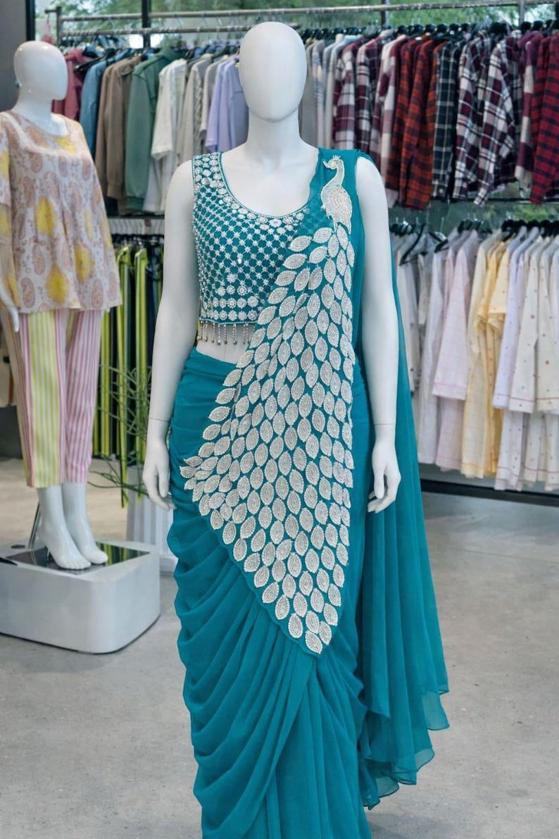 May include: A teal saree featuring a sleeveless blouse with intricate silver embroidery and a matching teal skirt with a prominent silver peacock feather pattern draped across the front. The ensemble includes a long, flowing teal fabric panel.