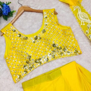 May include: A bright yellow sleeveless blouse featuring a white grid pattern and gold sequin embellishments, paired with a matching yellow skirt that has a white scalloped border with a gold sequin peacock design along the edge.