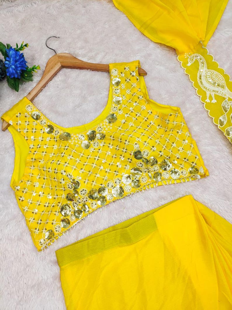 May include: A bright yellow sleeveless blouse featuring a white grid pattern and gold sequin embellishments, paired with a matching yellow skirt that has a white scalloped border with a gold sequin peacock design along the edge.