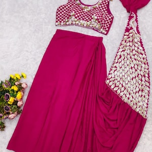 May include: A magenta two-piece outfit consisting of a sleeveless crop top and a long skirt. The top features a white lattice pattern with floral embellishments. The skirt includes a long, draped panel decorated with a large, white peacock motif.