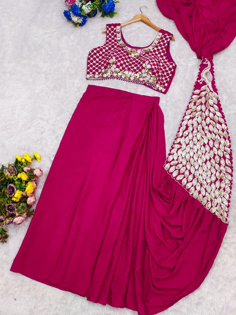 May include: A magenta two-piece outfit consisting of a sleeveless crop top and a long skirt. The top features a white lattice pattern with floral embellishments. The skirt includes a long, draped panel decorated with a large, white peacock motif.