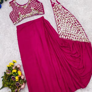 May include: A magenta two-piece outfit consisting of a sleeveless blouse and a skirt. The blouse features a white grid pattern with gold embellishments. The skirt includes a long, draped panel adorned with a large gold peacock motif and leaf-shaped sequins.