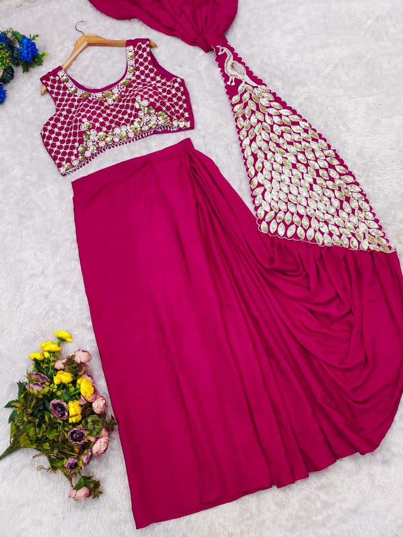 May include: A magenta two-piece outfit consisting of a sleeveless blouse and a skirt. The blouse features a white grid pattern with gold embellishments. The skirt includes a long, draped panel adorned with a large gold peacock motif and leaf-shaped sequins.