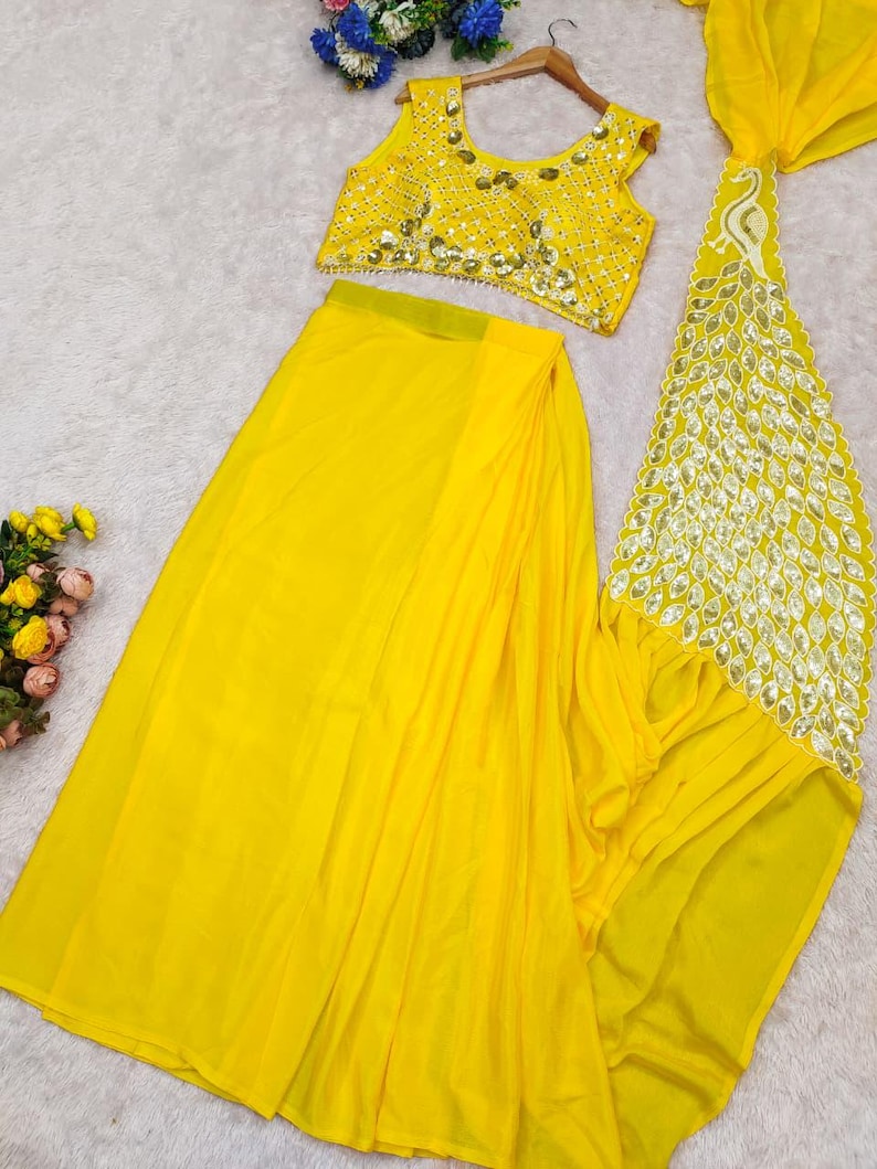 May include: A bright yellow two-piece outfit consisting of a sleeveless crop top and a long skirt. The top features intricate gold-toned embroidery and mirror-like embellishments. A matching yellow dupatta with a decorative peacock-shaped border.