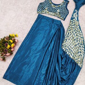 May include: A teal sleeveless crop top featuring a white grid pattern with gold embellishments, paired with a matching teal satin skirt that includes a long, flowing panel decorated with a gold peacock feather design and scalloped edges.