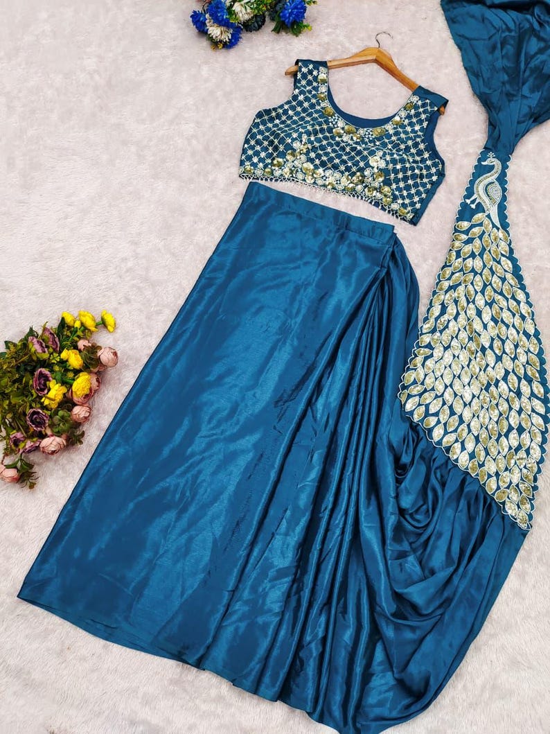 May include: A teal sleeveless crop top featuring a white grid pattern with gold embellishments, paired with a matching teal satin skirt that includes a long, flowing panel decorated with a gold peacock feather design and scalloped edges.