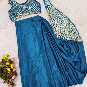 May include: A teal blue sleeveless blouse with a white lattice pattern and floral embroidery, paired with a matching teal blue skirt featuring a long, flowing panel decorated with a large white and gold peacock feather motif and sequins.